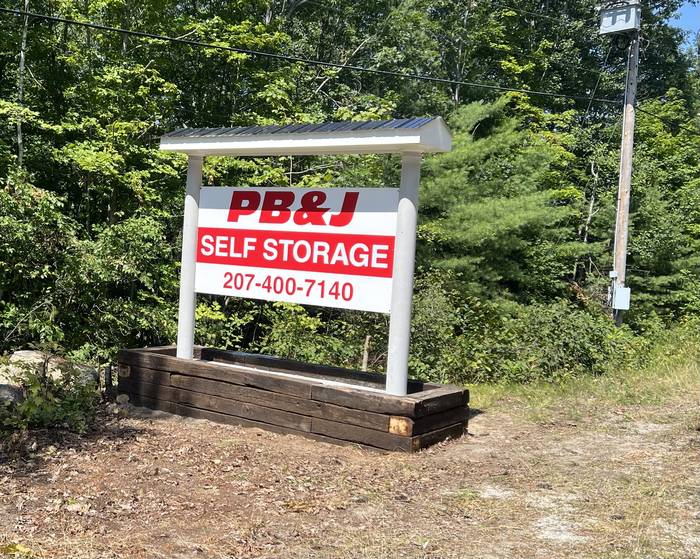 Big red and white PB&J Self Storage roadside sign with the phone number 207-400-7140, on white posts in a timber planter along Dole Ridge Road in Limerick, Maine